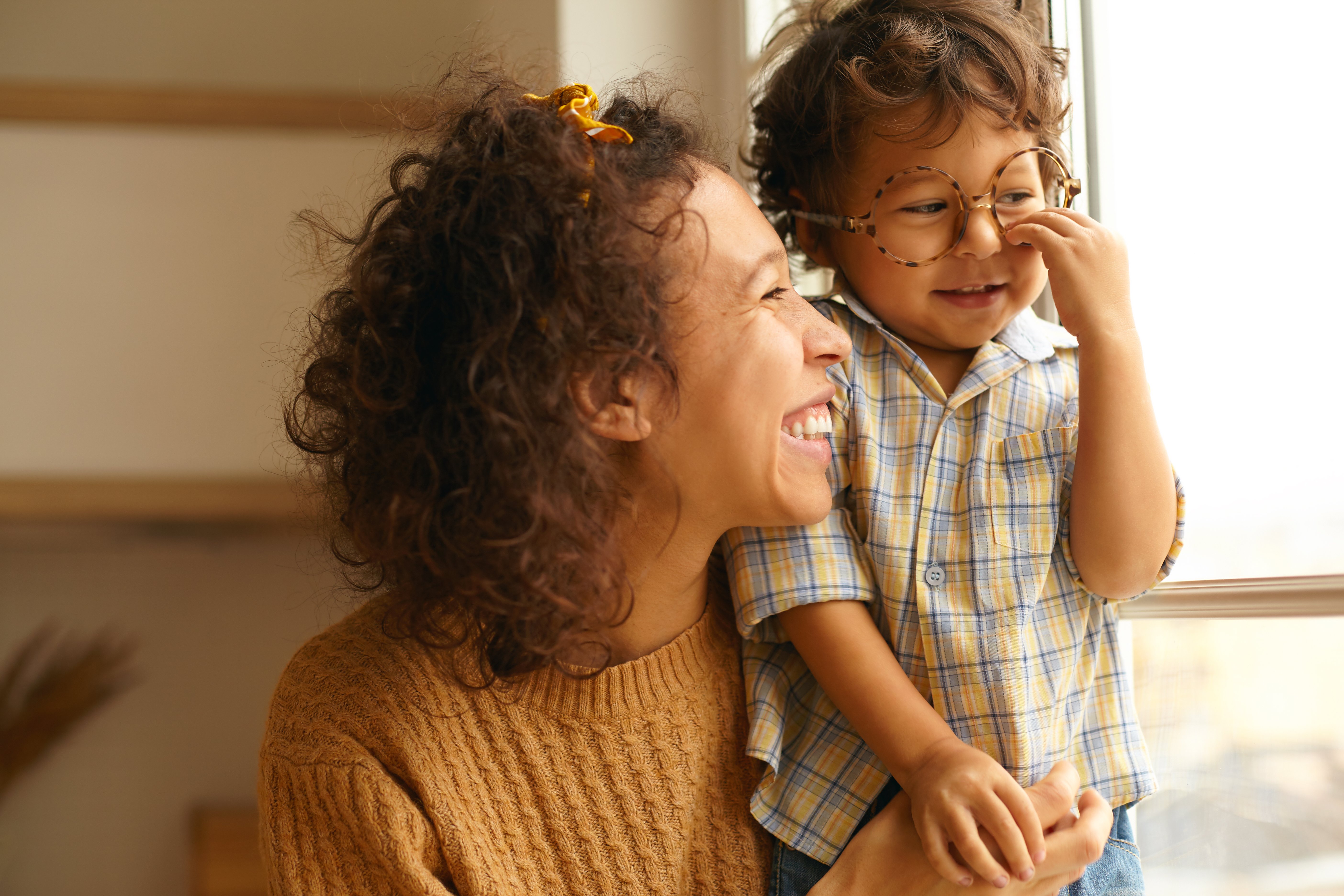 close-up-image-of-happy-young-wavy-haired-hispanic-female-posing-by-window-embracing-baby-son-cute-three-year-old-boy-wearing-round-eyeglasses-spending-day-at-home-family-and-relationships